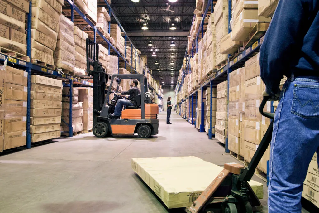 A warehouse worker operating a forklift among tall racks of palletized boxes.